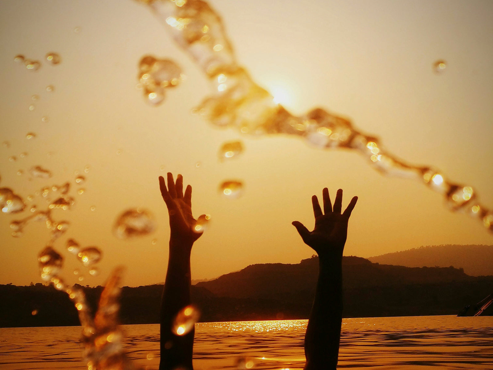 Silhouette of hands reaching out of water at sunset with droplets and golden sunlight