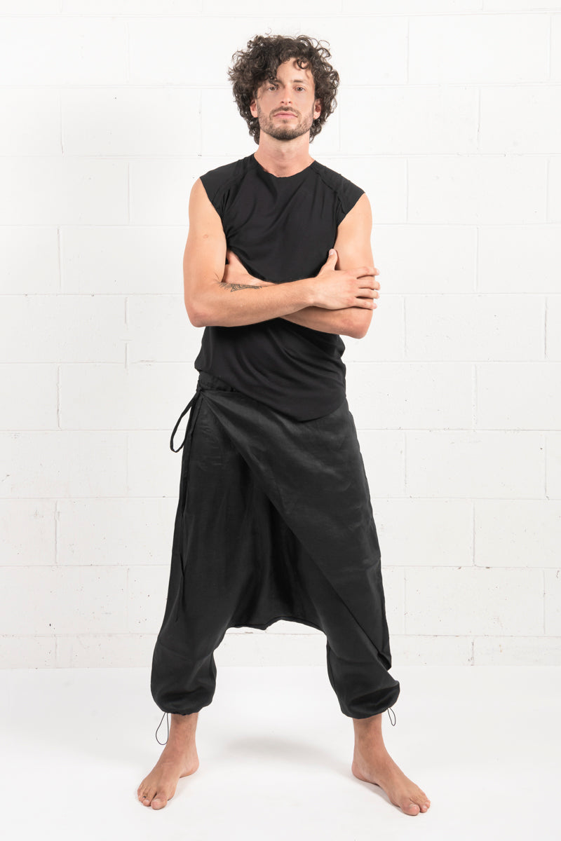 Man with curly hair in a black sleeveless top and draped trousers standing confidently against a white brick wall