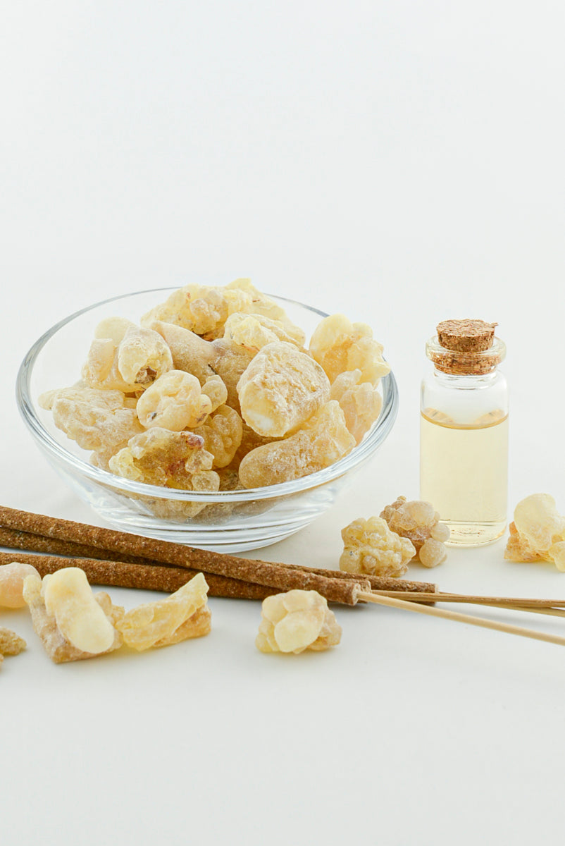 Glass bowl of natural frankincense resin next to a small bottle of essential oil and aromatic sticks on a white background