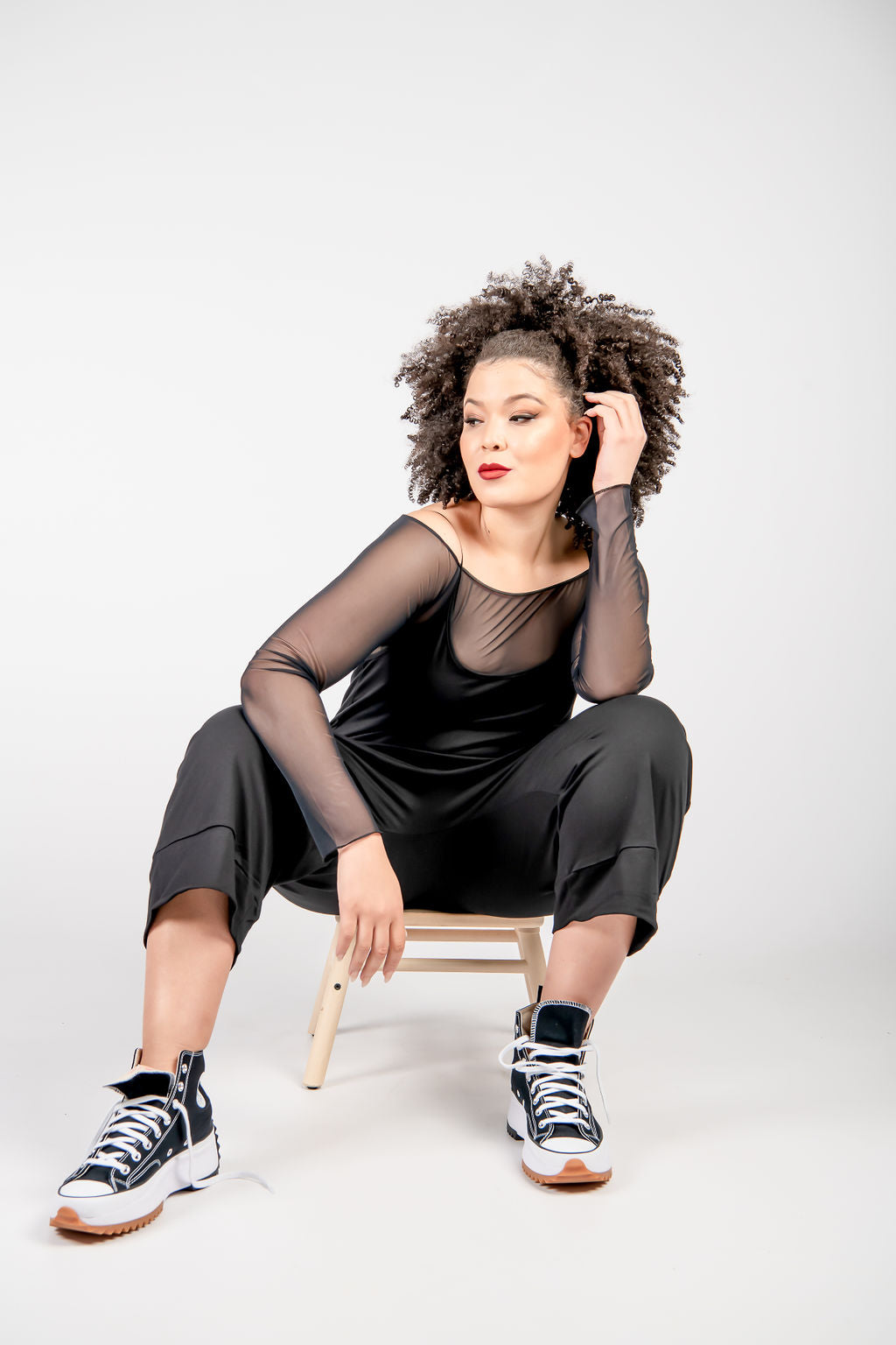 Fashionable young woman with curly hair seated on a stool, wearing a sheer black dress over a black outfit, paired with high-top sneakers