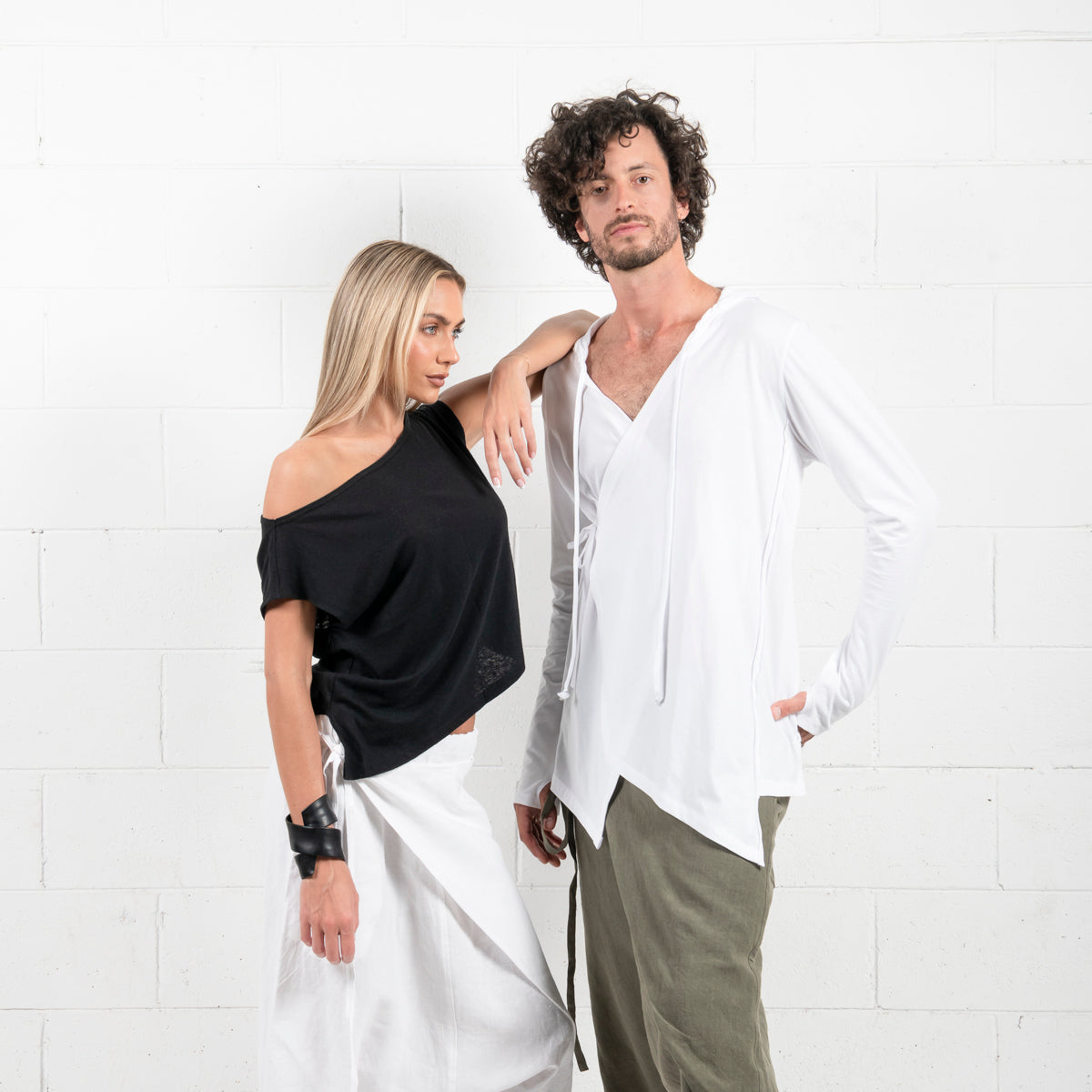 Fashionable couple posing in a studio, woman in a black off-shoulder top and white pants, man in a white hooded shirt and green trousers