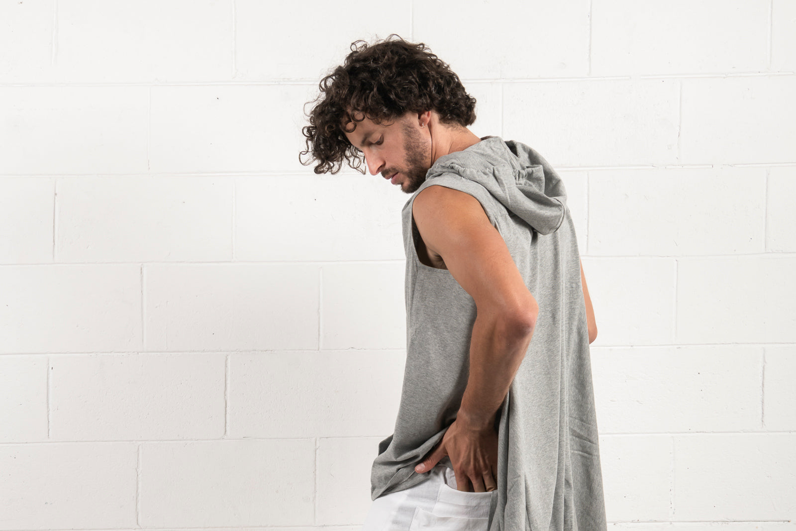 Man in a grey sleeveless hoodie and white pants standing against a white brick wall, looking downward