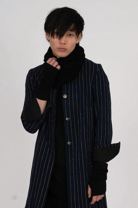 A model wearing a long navy blue pinstripe coat with black cashmere loop scarf  black gloves, loose black skirt, and black boots,
