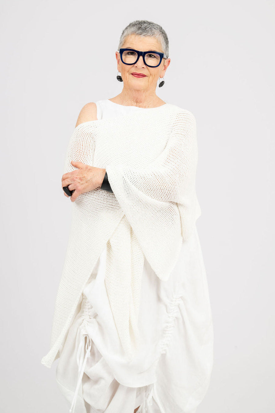 An older woman with short gray hair and black-rimmed glasses is wearing a white knit shrug and white draped clothing against a light gray studio background.