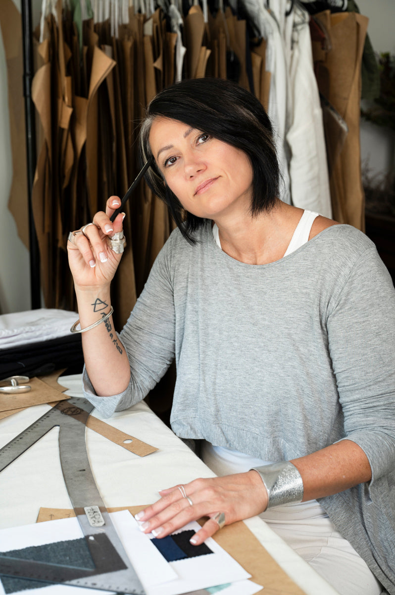 Fashion designer with short black hair holding a pen, sitting at her workspace surrounded by pattern papers and fabric samples