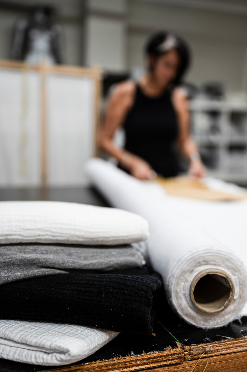 Focused shot of fabric rolls and textiles in the foreground with a blurred fashion designer working in the background in a studio
