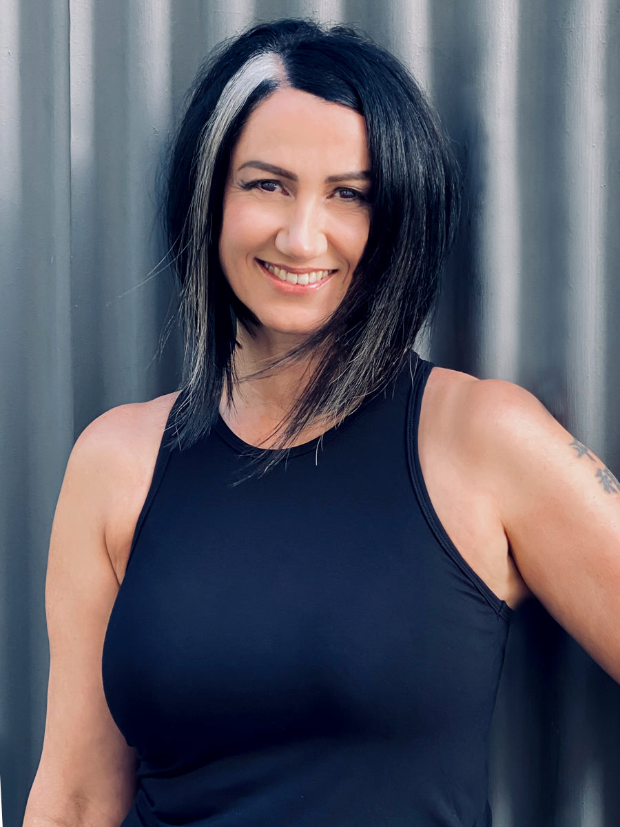 Portrait of a smiling woman with black hair and a white streak, wearing a tank top, standing against a grey corrugated background