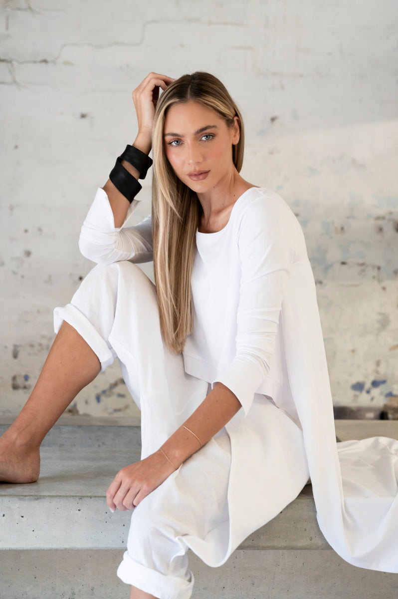 Elegant woman in a white outfit sitting on a concrete step, hand in hair, against a textured wall background
