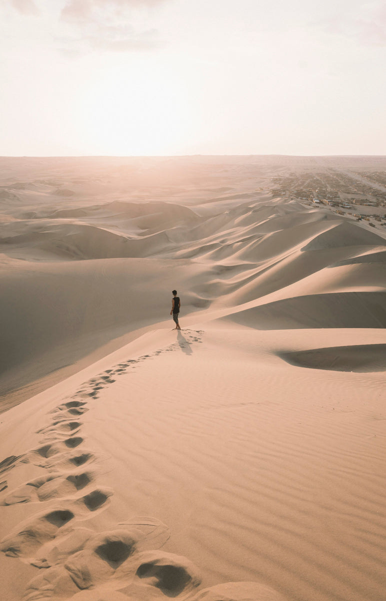 Lone figure walking on vast sand dunes, leaving a trail of footprints, with a soft sunset in the background